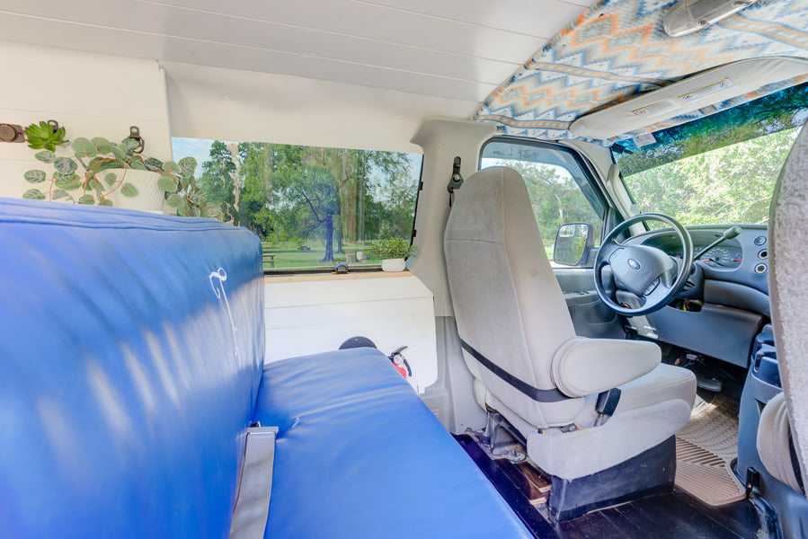 Converted van interior for RV rental: cozy blue seating, driver's cockpit, and serene park view from side window.