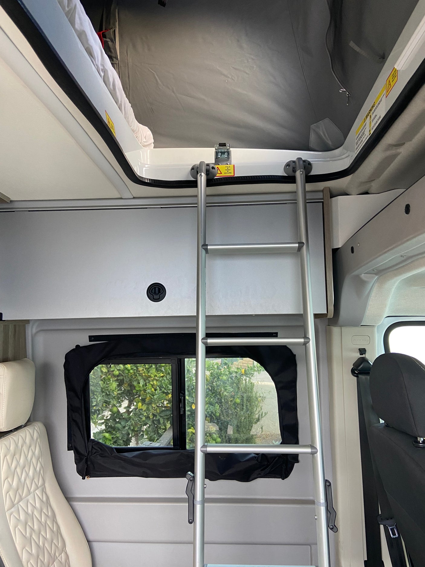 Inside an RV rental: a ladder to a pop-top bed, storage cabinets, and a window looking out to trees.