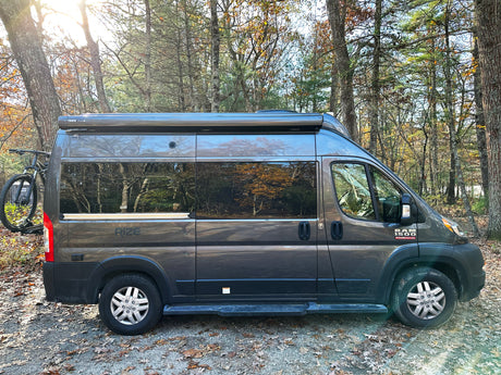 Explore nature with this grey Ram ProMaster camper van rv rental, featuring a bike rack, parked among sunlit trees.