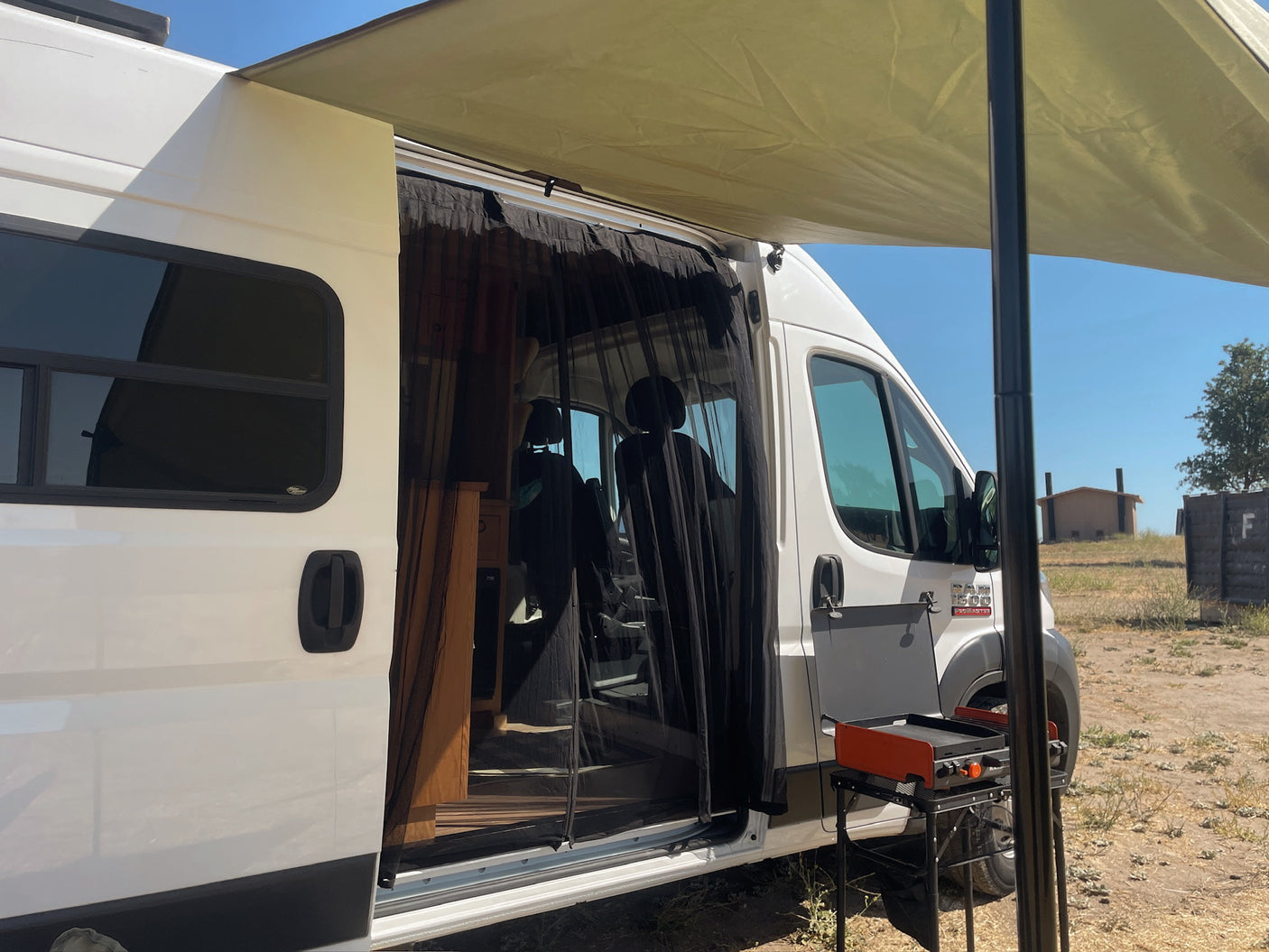 This RV rental features an open side door with a bug net, a deployed awning, and an outdoor grill setup for camping.