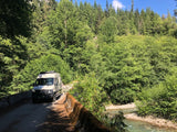 RV rental adventure: Camper van crossing a mossy bridge over a clear river, surrounded by a lush forest under a blue sky.