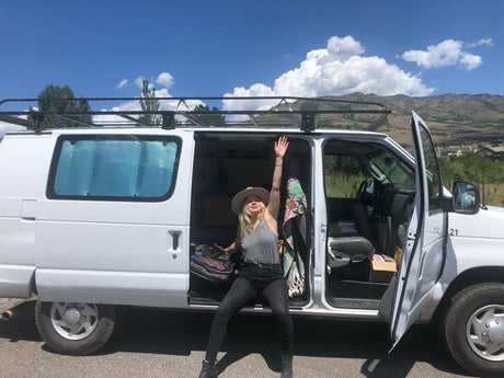 Woman waves from her white RV rental van, ready for mountain adventure. Scenic travel under blue sky.