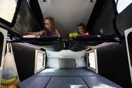 RV rental fun! Two girls enjoy the upper bunk of a family-friendly camper interior. Lower bed and window visible.