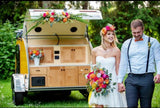 A happy couple holds hands by a yellow teardrop trailer with an open kitchen, decorated with flowers for their wedding. Consider this unique RV rental!