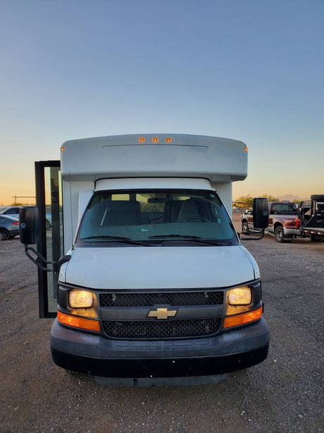 White Chevrolet cutaway bus, perfect for RV rental. Front view with lights on, parked on a gravel lot.
