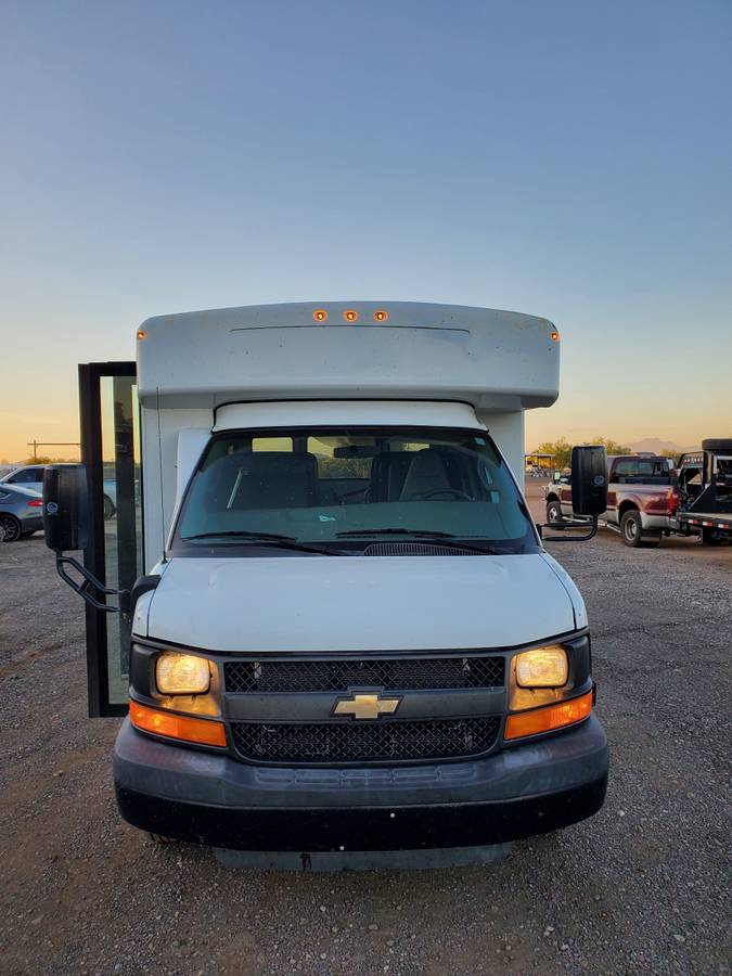 White Chevrolet cutaway bus, perfect for RV rental. Front view with lights on, parked on a gravel lot. — photo 6