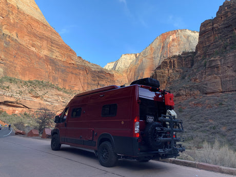 Maroon RV rental van parked on a road, flanked by majestic sandstone cliffs under a clear blue sky, ideal for scenic adventures.