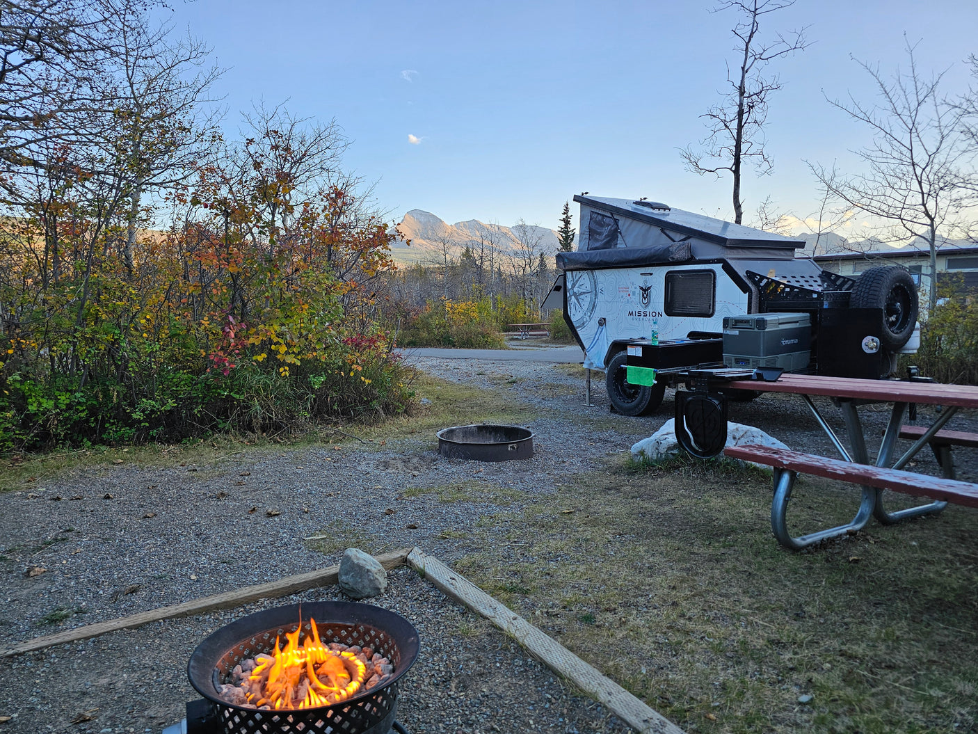 An overland trailer RV rental at a scenic fall campsite, featuring a warm fire pit, picnic table, and majestic mountains in the background.