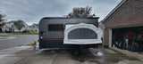 This dark gray travel trailer, an RV rental with a rear pop-out tent, is parked on a driveway, ready for adventure or a camping trip.