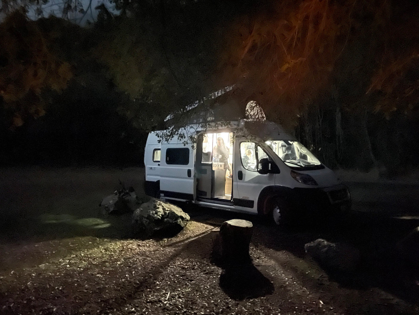 A bright white rv rental campervan is parked at night in a dark forest, its open door revealing two people within.
