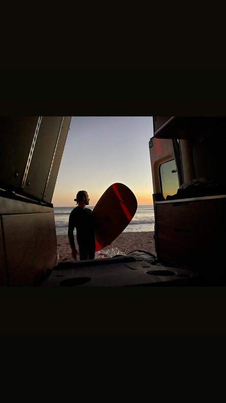 Surfer with board at sunset on beach, viewed from inside an RV rental. Ocean waves and colorful sky.