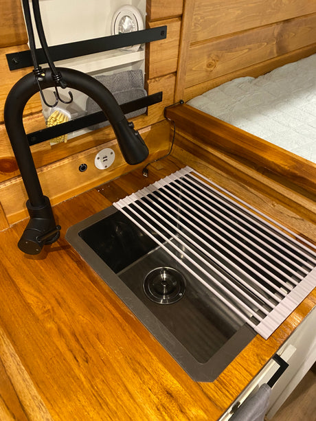 RV rental interior: cozy kitchen with wooden counter, sink, black faucet & drying rack. USB port and bed nearby.