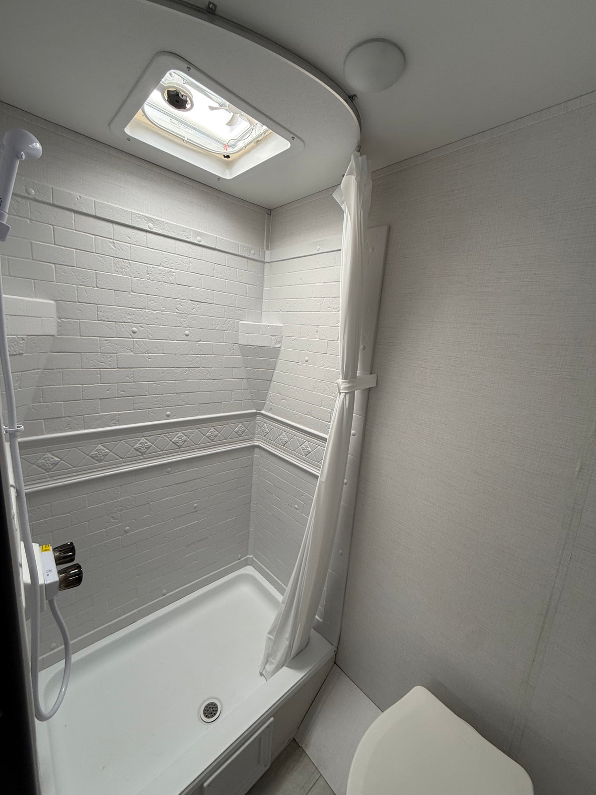Compact RV rental bathroom: white shower stall with textured brick-look walls, overhead vent, shower curtain, and adjacent toilet. — photo 12