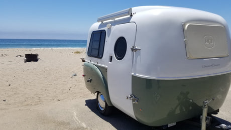 A stylish, two-tone Happier Camper at a serene beach. This RV rental offers scenic ocean travel for outdoor enthusiasts.
