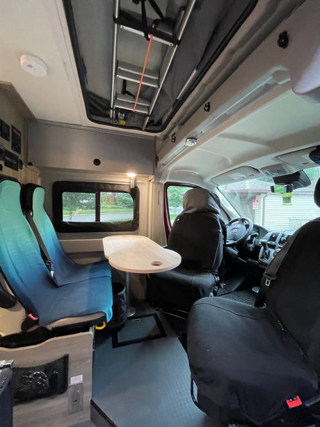 RV rental interior: Two rear seats with blue covers, a small table, and an overhead ladder. Driver/passenger seats visible.