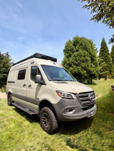This light-colored Winnebago Revel RV rental, a Mercedes Sprinter camper van, is parked on grass surrounded by pine trees.