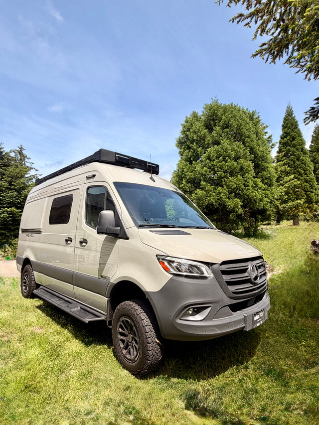 This light-colored Winnebago Revel RV rental, a Mercedes Sprinter camper van, is parked on grass surrounded by pine trees.