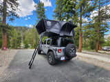 Experience nature with this gray Jeep Wrangler rv rental featuring a rooftop tent, set in a mountain campground among tall pines.