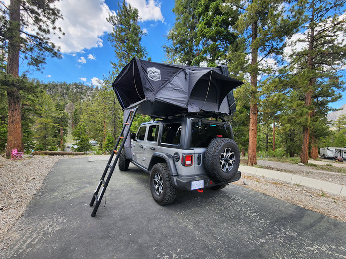 Experience nature with this gray Jeep Wrangler rv rental featuring a rooftop tent, set in a mountain campground among tall pines.