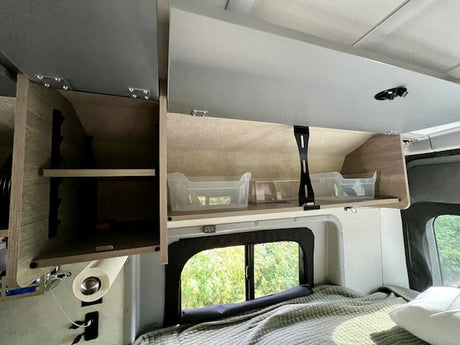 Inside an RV rental: open overhead cabinets with clear bins, shelves, a window, and a bed. Functional space.