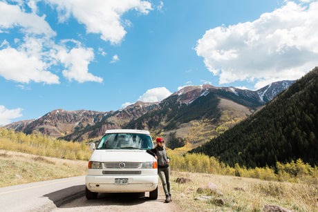 A person poses with a white rv rental van on a mountain road, surrounded by vibrant autumn trees & snow-capped peaks under a blue sky.