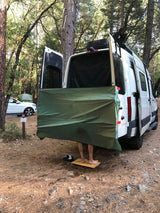 A person uses an outdoor shower behind an RV rental in a forest campsite, standing on a wooden mat.