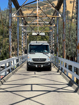 A light gray RV rental van crossing a historic truss bridge, surrounded by scenic hills. Ideal for travel adventures.