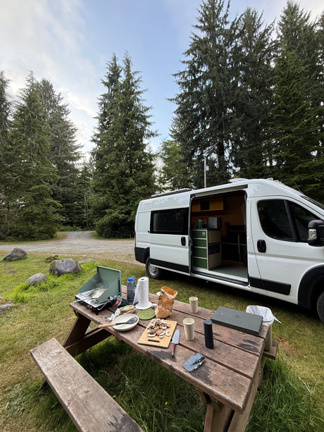 RV rental camping: cooking outdoors on a picnic table next to a white camper van at a forest campsite.