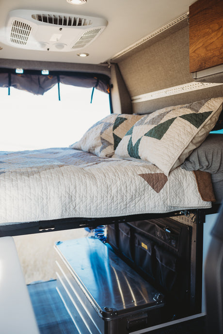 Cozy RV rental interior: elevated bed with quilted bedding, ceiling AC, and practical under-bed storage.