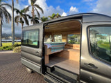 Experience van life! This grey RV rental features a cozy wood-paneled interior with a bed, ready for your tropical road trip adventure.