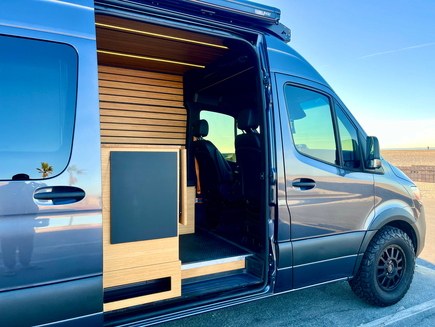 Spacious RV rental van with a sleek blue exterior, modern wood interior, and off-road tires, parked by a sunny beach.