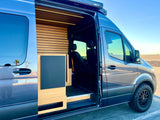 Spacious RV rental van with a sleek blue exterior, modern wood interior, and off-road tires, parked by a sunny beach.