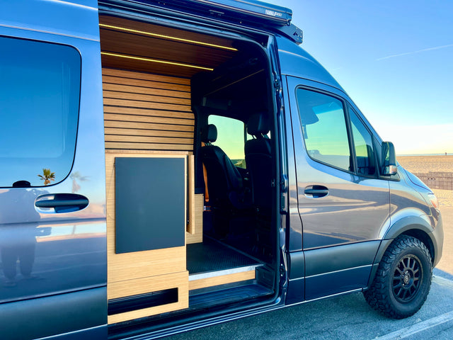 Spacious RV rental van with a sleek blue exterior, modern wood interior, and off-road tires, parked by a sunny beach.