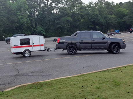 Black truck towing a white teardrop RV rental camper, parked on asphalt with trees in the background.