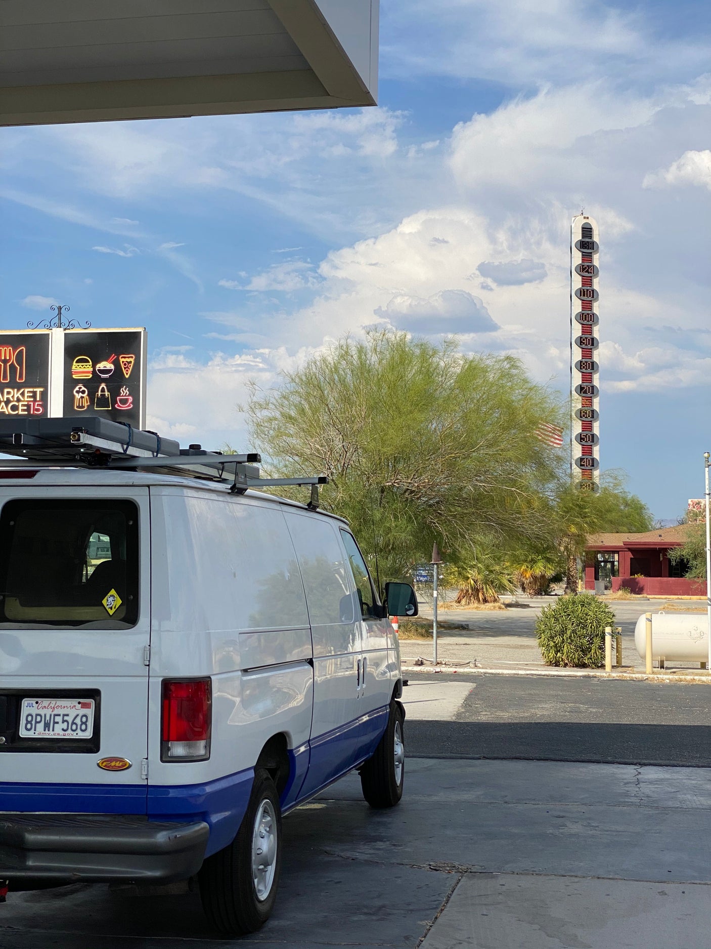 An RV rental van with CA plates, parked by a tall desert thermometer showing extreme heat and a market sign under a cloudy sky.