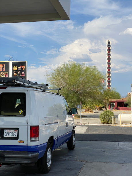 An RV rental van with CA plates, parked by a tall desert thermometer showing extreme heat and a market sign under a cloudy sky.