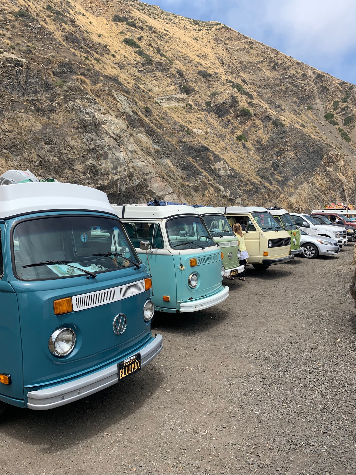 A row of colorful vintage Volkswagen camper vans parked on a gravel lot beside a rugged hill, suggesting fun RV rental options.