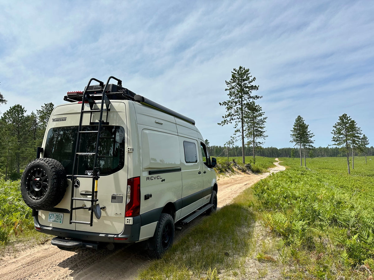  offering breathtaking views under a cloudy sky.|Adventure awaits! This cream Winnebago Revel **rv rental** navigates a sandy track through a vibrant green forest.|RV rental camper van parked by a scenic lake — photo 2