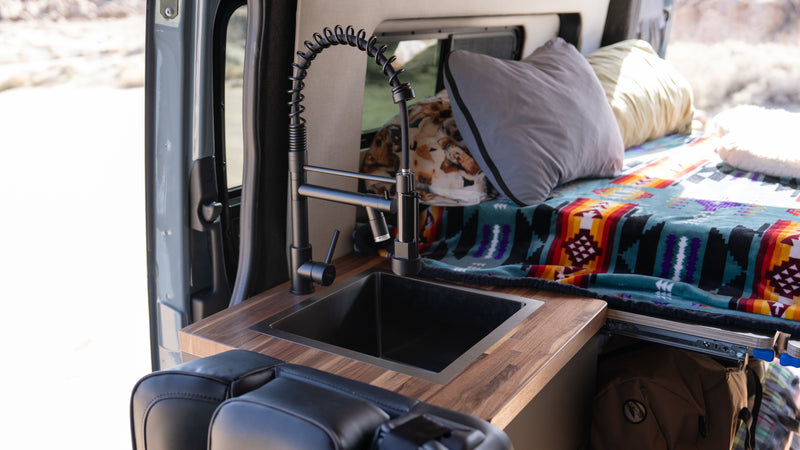 Inside an RV rental: a sleek black sink & wood counter, alongside a cozy bed with colorful blankets, ideal for van life travel. — photo 4