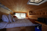 Inside a cozy RV rental: a wooden-paneled camper van bedroom with a comfortable bed, pillows, and snowy window views.