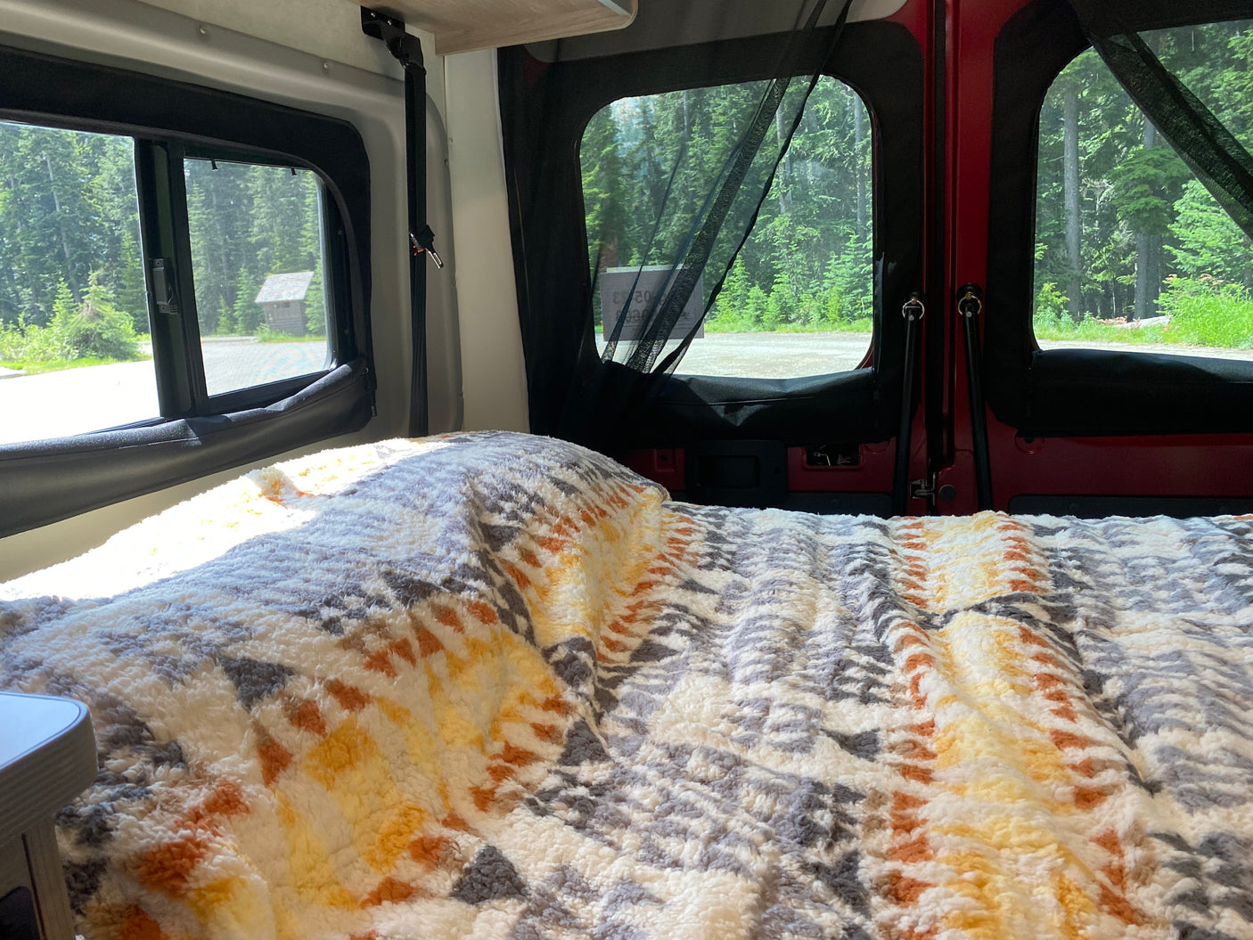Cozy RV rental interior with a comfy bed, offering scenic views of a dense forest and a small cabin.