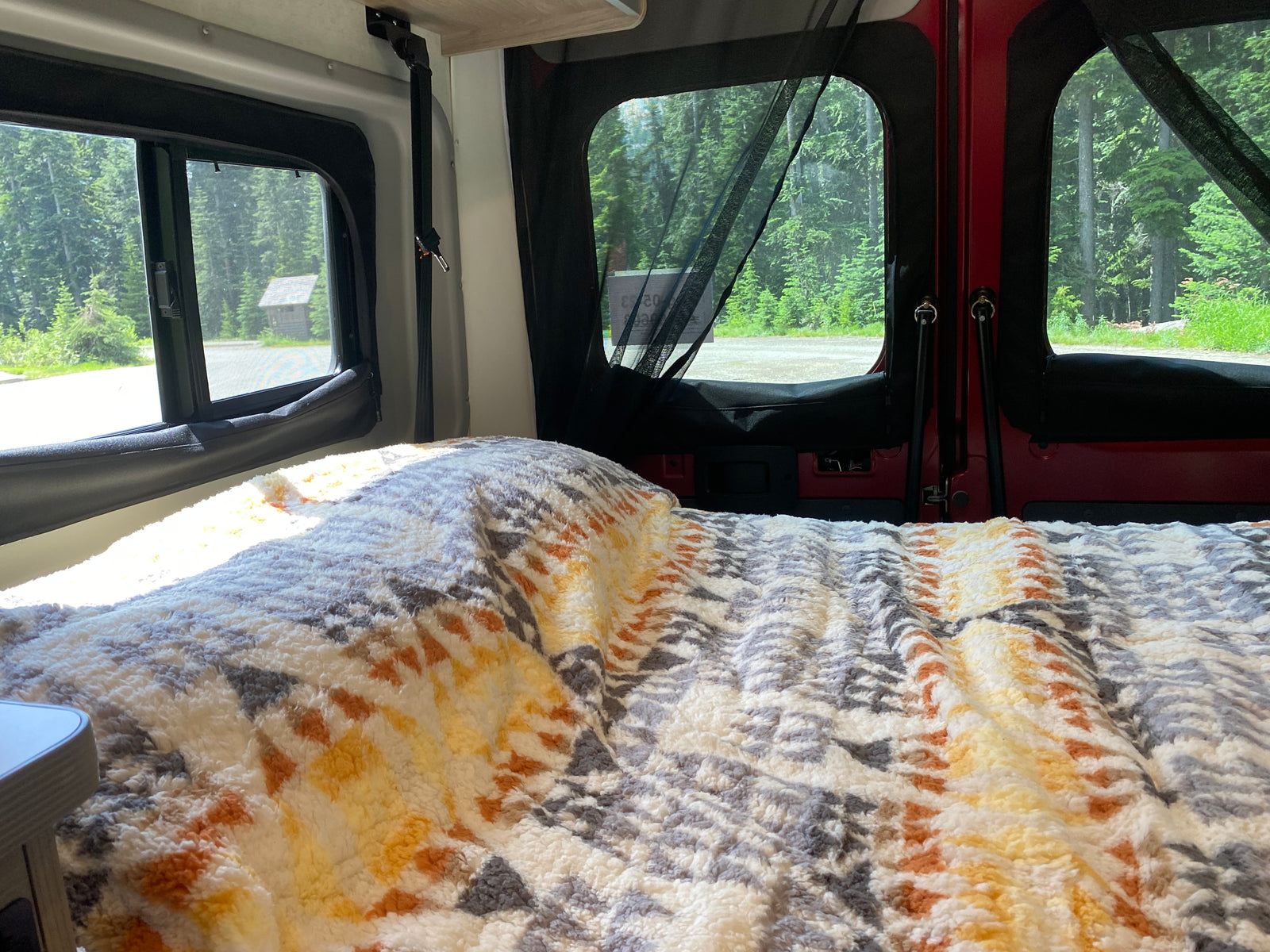 Cozy RV rental interior with a comfy bed, offering scenic views of a dense forest and a small cabin.