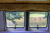 Inside an RV rental, two windows frame a peaceful view of a river, with lush trees on one bank and a green field on the other.