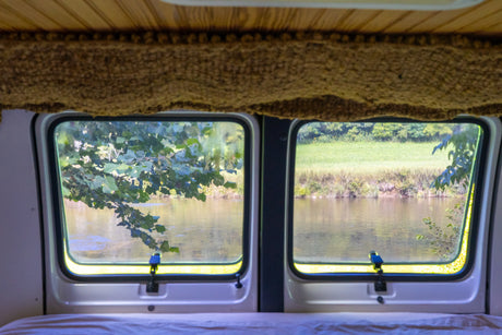 Inside an RV rental, two windows frame a peaceful view of a river, with lush trees on one bank and a green field on the other.