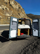 This RV rental van, with its cozy bed revealed, is parked on a scenic mountain road. Perfect for travel adventures!