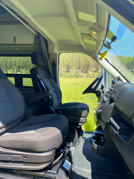 Interior of an rv rental parked in a lush green field, with front seats and steering wheel visible, overlooking a forest.