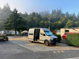 A light grey RV rental campervan parked at a scenic campground campsite 221, with misty evergreen trees and other RVs.