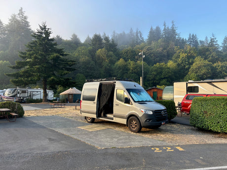 A light grey RV rental campervan parked at a scenic campground campsite 221, with misty evergreen trees and other RVs.