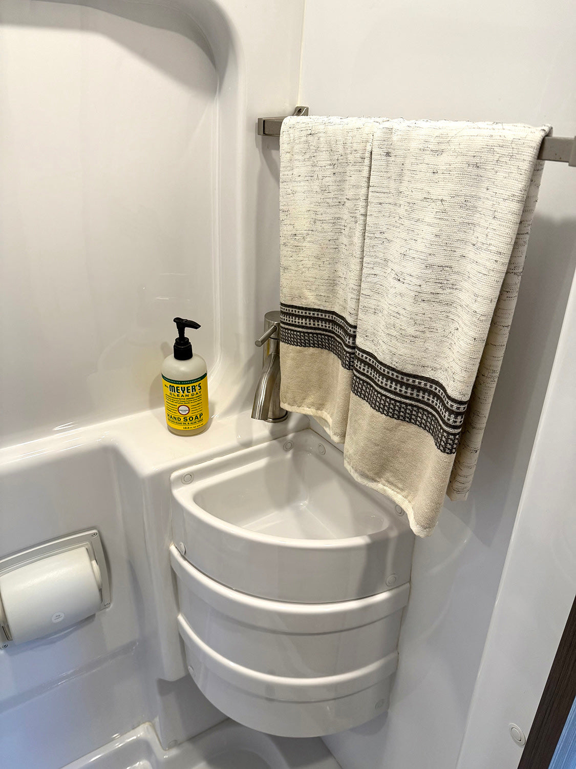 Compact RV rental bathroom featuring a corner sink, hand soap, toilet paper holder, and towel bar with a patterned towel.