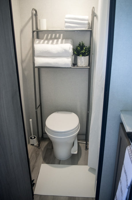 This RV rental bathroom features a compact toilet and over-toilet shelving with fresh towels and greenery. Essential travel amenities.